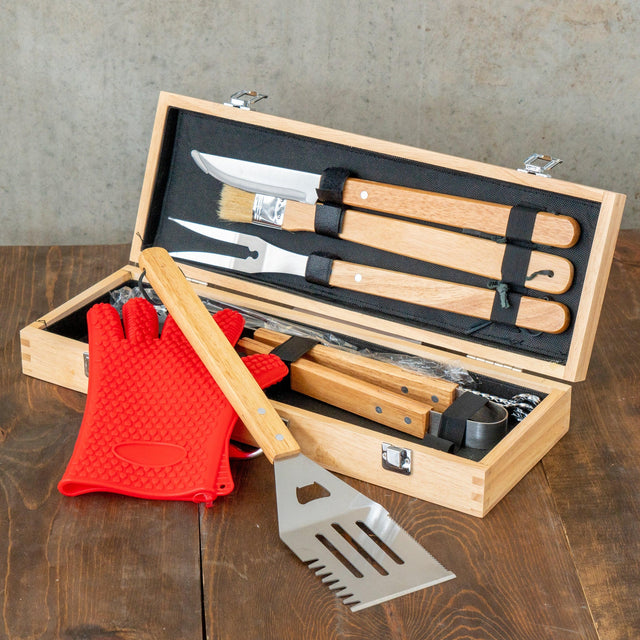Open wooden barbecue tool case with utensils and red grilling glove on a table.