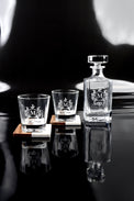 Engraved whiskey decanter and two rocks glasses on a dark reflective surface.