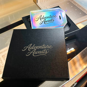Holographic Laserette luggage tag engraved with Adventure Awaits in a black gift box.